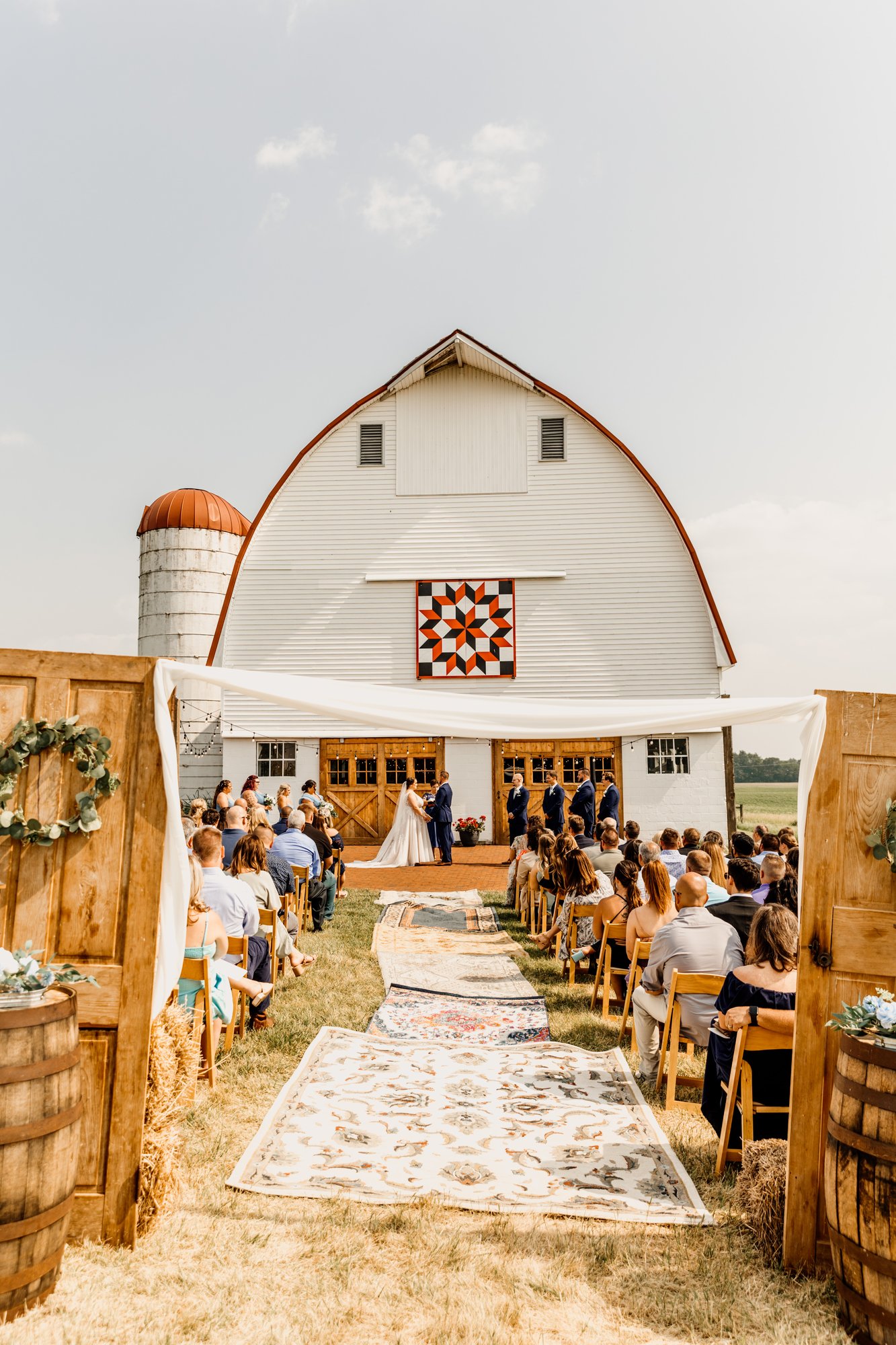 Ceremony on the brick patio in front of the white barn, vintage rugs lining the aisle.