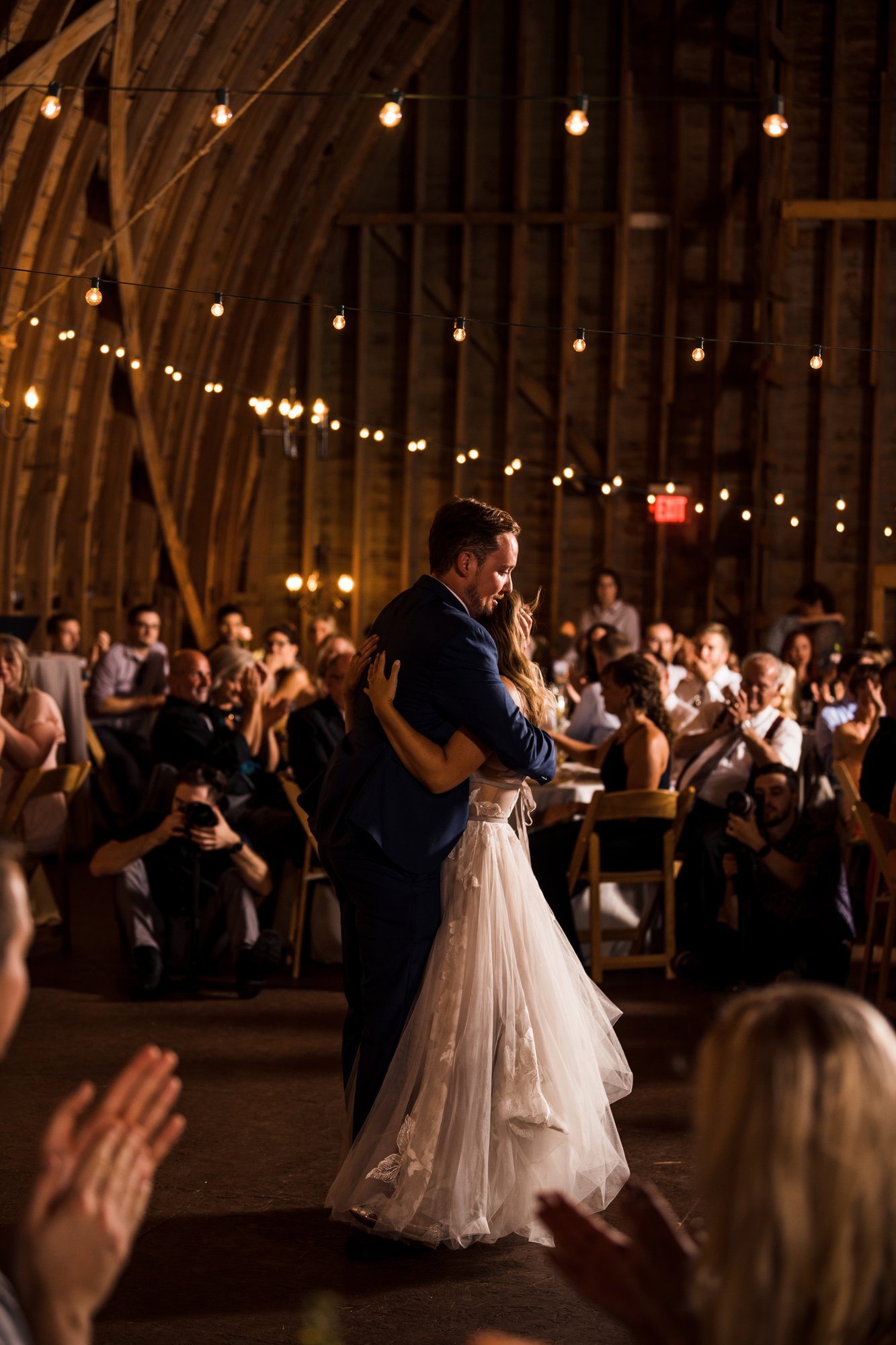 An embrace on the dance floor under string lights while guests applaud.