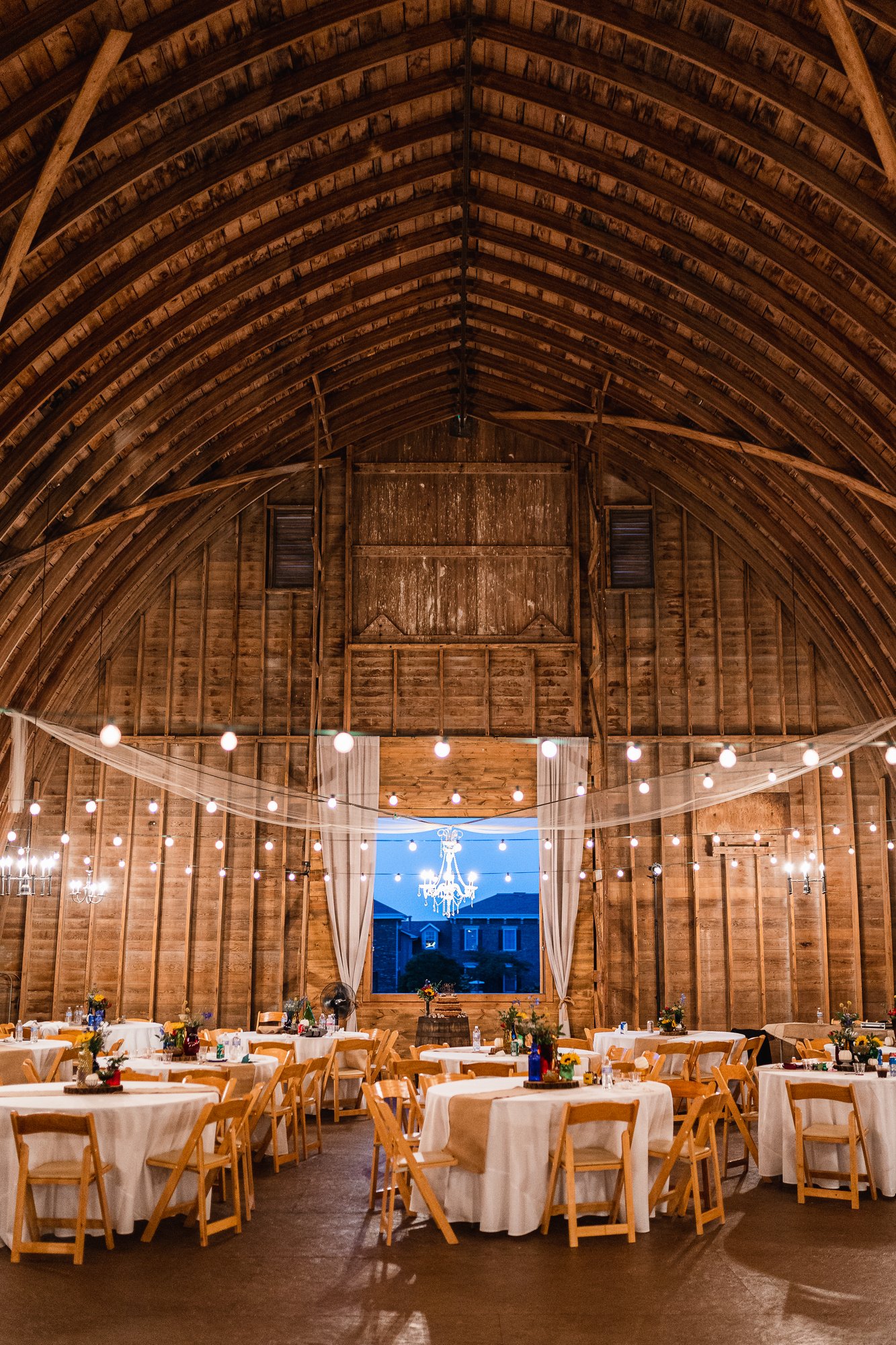 The barn at dusk set with round tables, string lights, and a chandelier hung in the window opening.