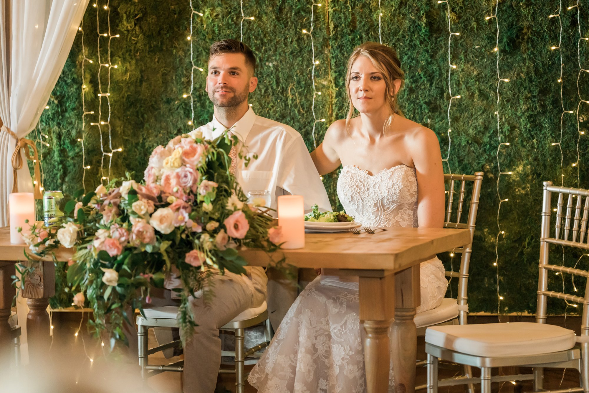 Couple seated at a wooden sweetheart table in front of a moss wall strung with lights.