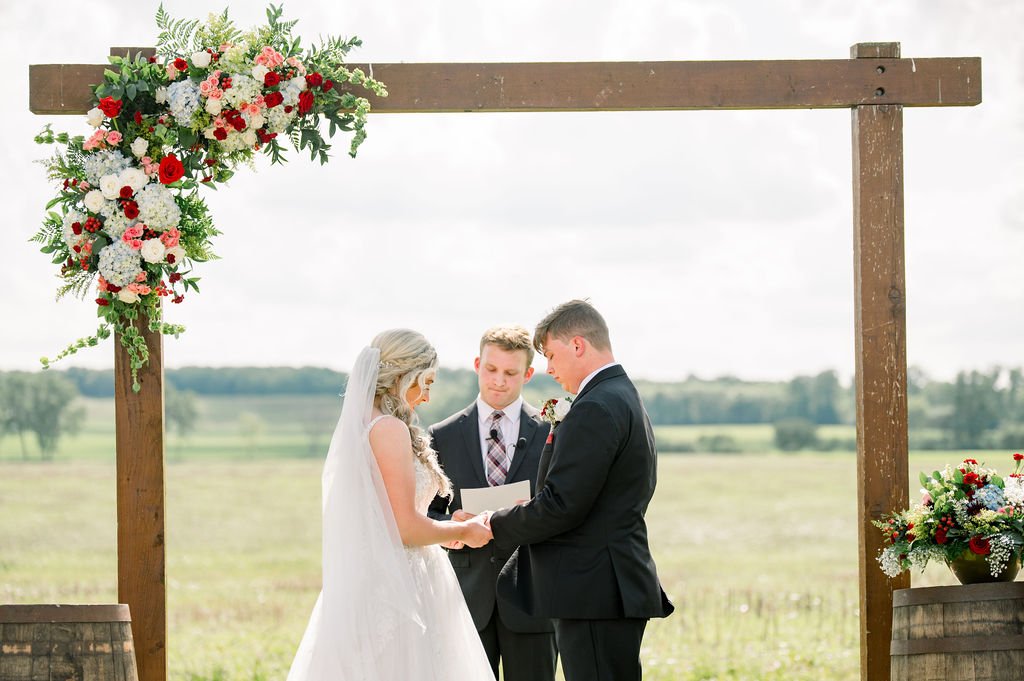 Bride and groom at the wooden arch with red and white florals in the open field.
