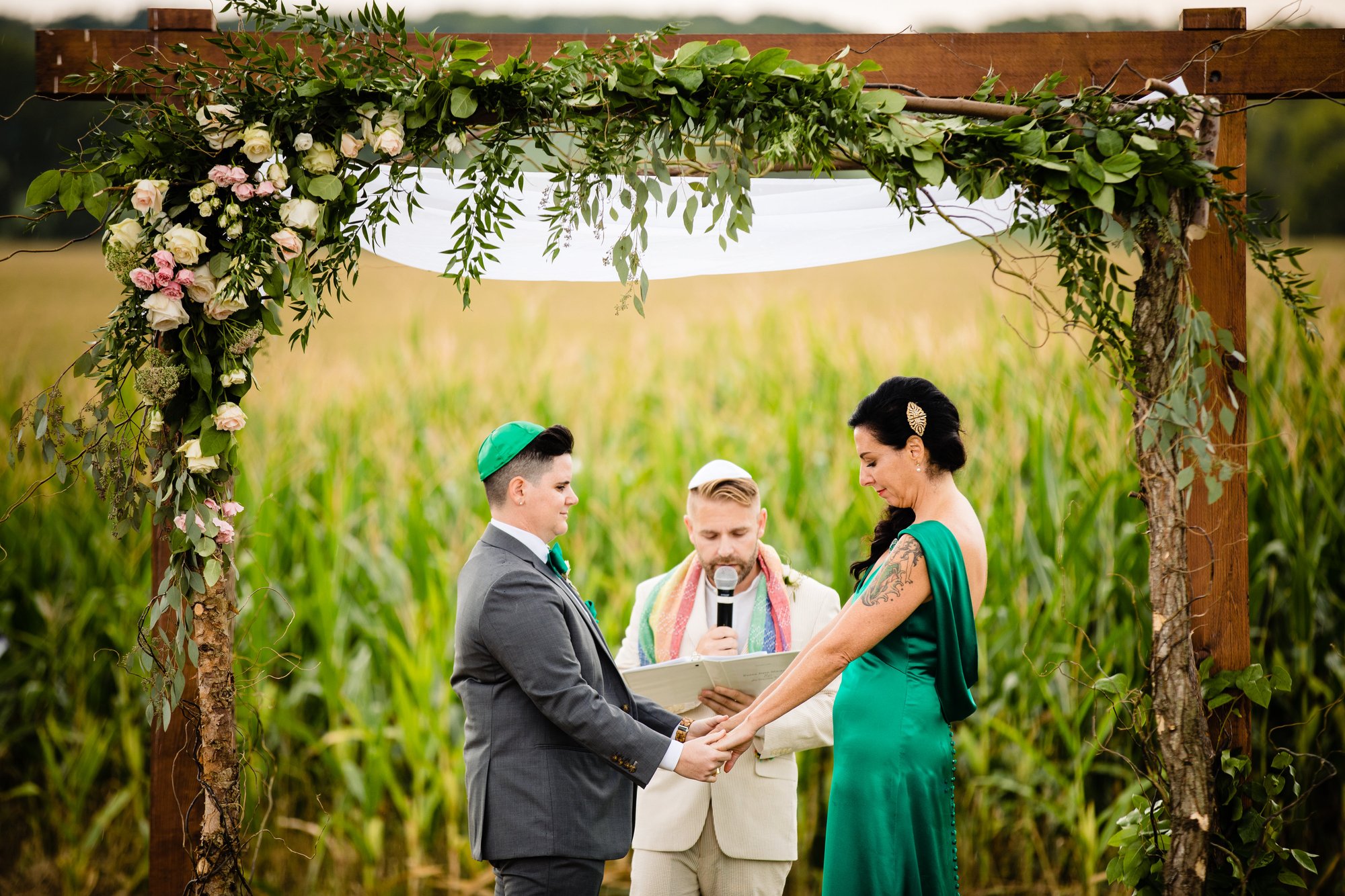 Two partners hold hands beneath the draped ceremony arch, cornfield behind them.