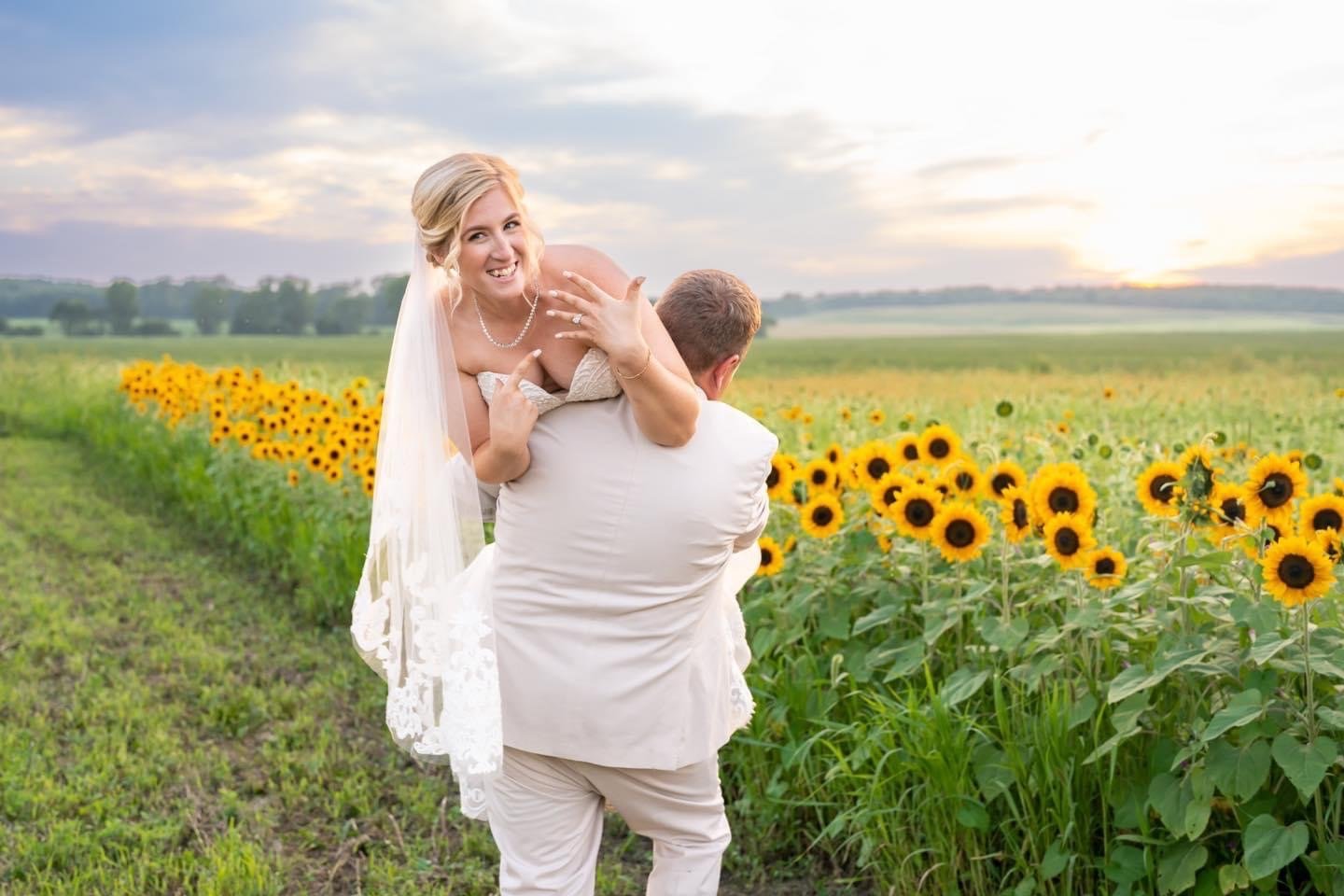 Groom carries the bride over his shoulder along the sunflower field while she laughs and shows her ring.