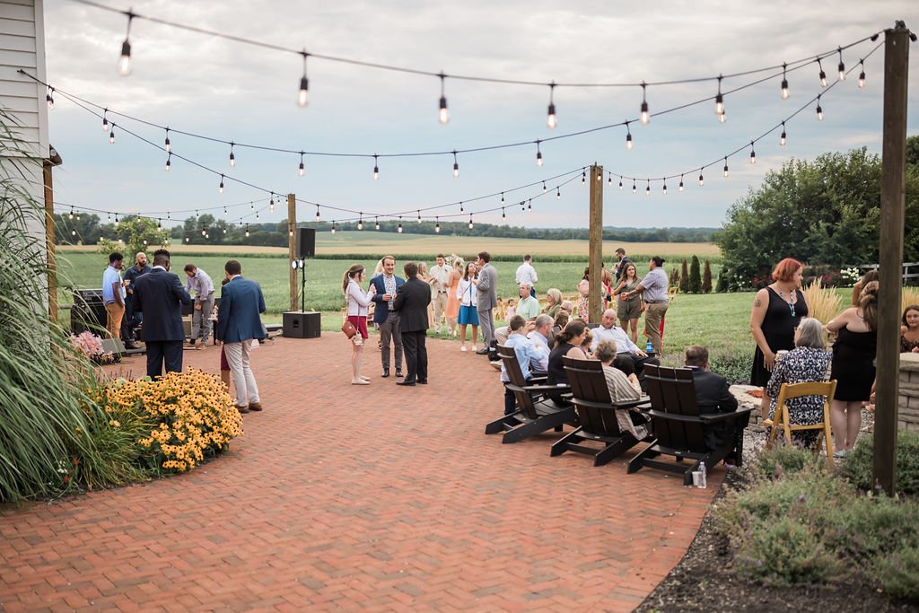 Guests mingle on the brick patio under cafe lights with fields beyond.